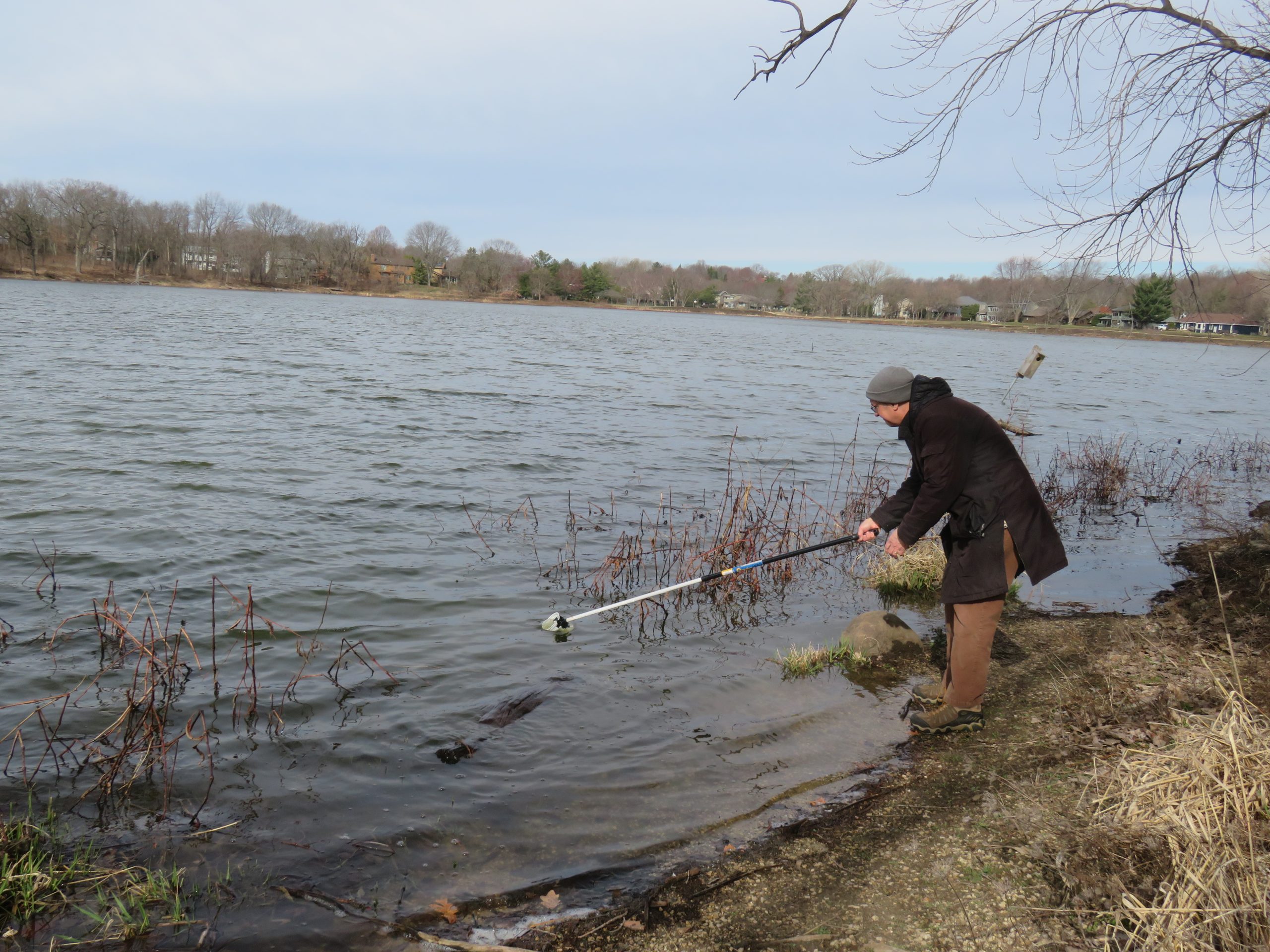 A man uses a water sampling tool to collect water from the edge of a pond in the wintertime.